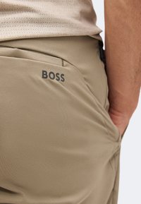 Κοντινή λήψη τσέπης μπεζ παντελονιού με το λογότυπο "BOSS" τυπωμένο πάνω από την τσέπη στη δεξιά πίσω πλευρά, που φοριέται από ένα άτομο.