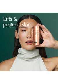 Femme à la peau lisse tenant le sérum Weleda Cell Longevity près de son visage sur un fond vert avec le texte "Soulève et protège la peau."