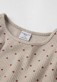 Beige ribbet top med små røde hjerte mønstre, rund halsudskæring og et label, der angiver mærke og størrelse.