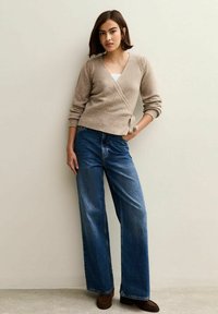 Beige Wickelpullover mit langen Ärmeln, kombiniert mit weit geschnittenen blauen Jeans. Das Outfit beinhaltet braune Mokassins. Leichte Textur.