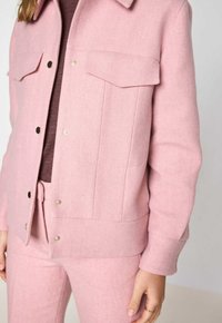 Veste en mélange de laine rose au design droit, dotée d'un col pointu, de deux poches poitrine et de fermetures à boutons-pression. Texture douce avec un motif subtil.