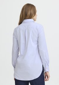 Camicia a maniche lunghe di un leggero viola, con colletto a bottoni, dettagli con piega sul retro e orlo arrotondato. Il tessuto sembra morbido e liscio.