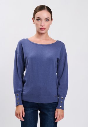 Vrouw met donker haar in een lage knot, draagt een blauwe trui met lange mouwen met knoopdetails bij de manchetten en donkerblauwe spijkerbroek.