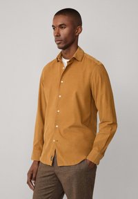 Camisa de cordón en color amarillo mostaza, de manga larga, con botones en la parte delantera y una textura suave. Combinada con pantalones de cuadros marrones.