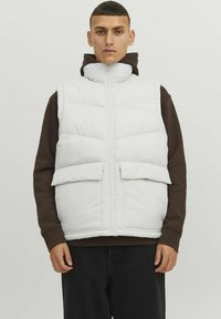 White puffer vest with a high collar, front zip, and two large flap pockets. Worn over a brown hoodie, featuring a smooth, padded texture.