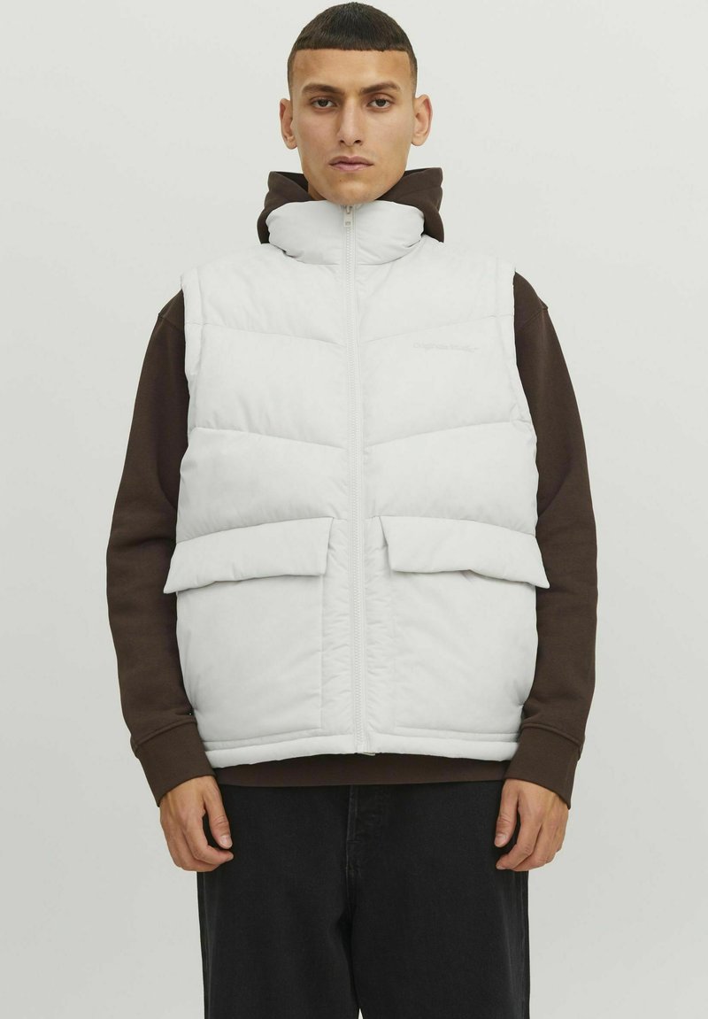 White puffer vest with a high collar, front zip, and two large flap pockets. Worn over a brown hoodie, featuring a smooth, padded texture.