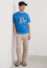 Tricou albastru cu grafică de barcă, pantaloni din in bej și sandale maro. Bărbat sprijinit de un perete, purtând un capac bleumarin.