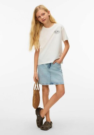 Young girl with long blonde hair wearing white printed t-shirt, denim skirt, brown clogs, and holding woven handbag.