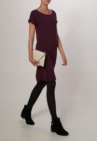 Abito in maglia viola a maniche corte, indossato con collant neri e stivaletti neri alla caviglia. Tiene una pochette beige con una patta triangolare. Texture liscia.