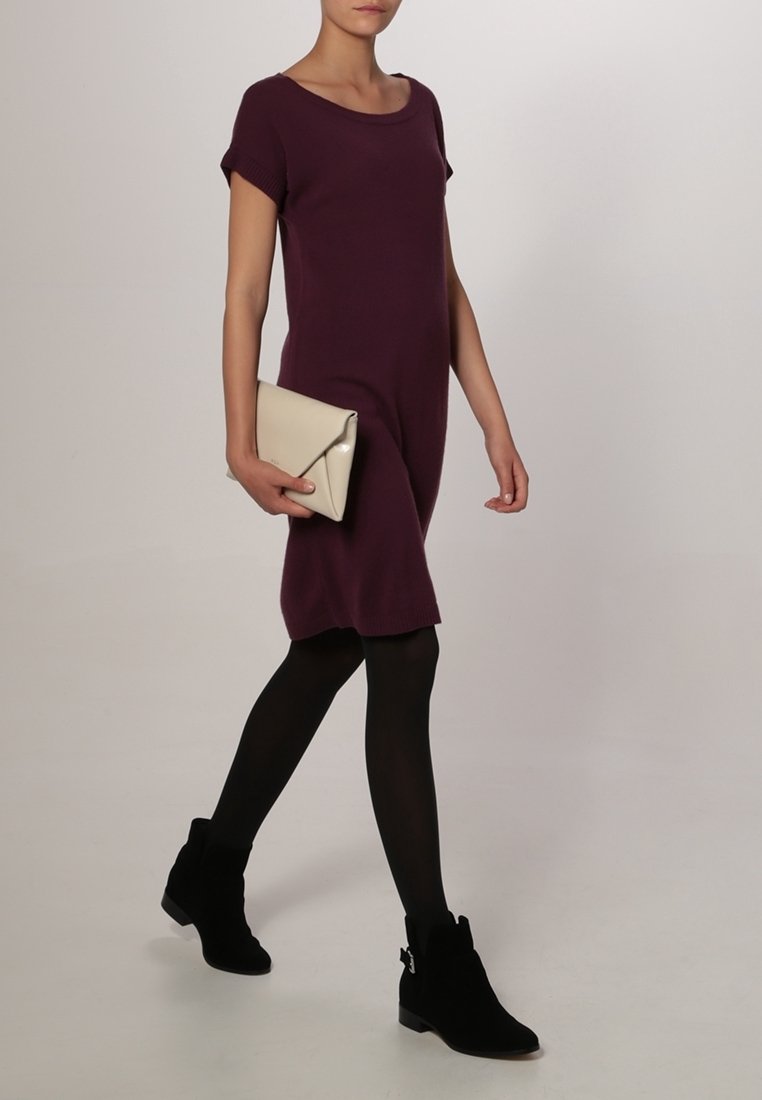 Abito in maglia viola a maniche corte, indossato con collant neri e stivaletti neri alla caviglia. Tiene una pochette beige con una patta triangolare. Texture liscia.