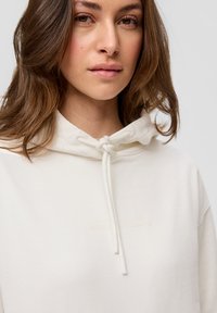 Jasno beżowa bluza z kapturem z zapięciem na sznurek i subtelnym nadrukiem współrzędnych geograficznych w dopasowanym kolorze na przodzie. Miękka faktura materiału.