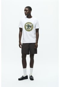 White t-shirt with a circular, colorful graphic design, paired with brown shorts, black loafers, and white ribbed socks.