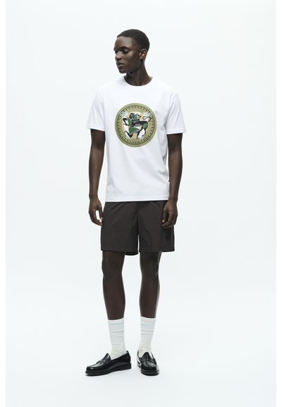 Tricou alb cu un design grafic circular și colorat, asortat cu pantaloni scurți maro, mocasini negri și șosete albe cu dungi.