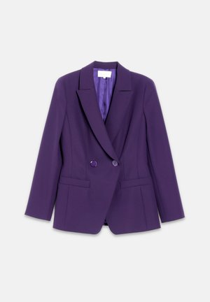 Kvinde i lilla blazer og plisseret nederdel med en blomstret bluse, sorte knæhøje støvler og en sort skuldertaske, stående selvsikkert.