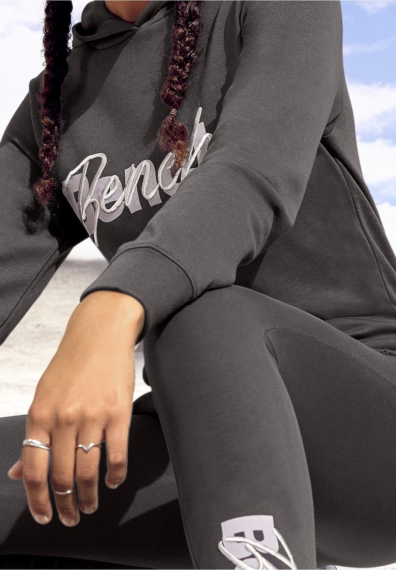 Sweat à capuche gris avec le logo "Bench" en blanc et leggings gris assortis. Coiffure tressée et plusieurs bagues en argent à la main. Texture du tissu douce.