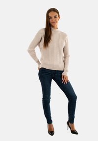 Maglione lavorato a maglia beige chiaro con motivo a texture, polsini a coste e colletto alto, abbinato a jeans slim fit blu scuro e tacchi neri.