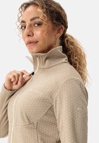 Giacca beige con zip, caratterizzata da un motivo testurizzato e un colletto alto. Presenta un logo sul petto e un cordino sulla zip.