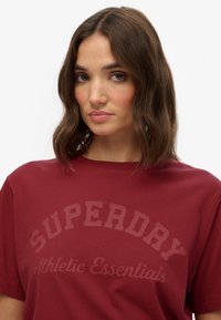 Bordeaux katoen t-shirt met een ronde halslijn en korte mouwen, met "SUPERDAY" en "Athletic Essentials" in contrasterende druk.