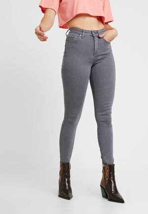 Vrouw met grijze high-waisted skinny jeans, een kort roze topje en enkellaarsjes met een puntige neus in slangenhuidpatroon, staand met één hand in de zak.