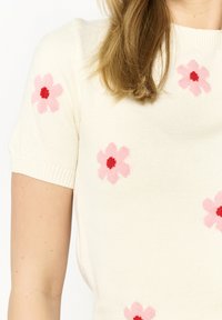 Knit sweater in cream with short sleeves, featuring pink flowers with red centers, creating a floral pattern throughout the fabric.