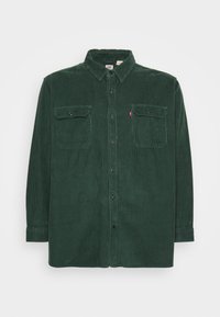 Chemise en velours côtelé vert foncé avec un col boutonné, des manches longues et deux poches avant. Présente une texture nervurée verticale et des boutons noirs.