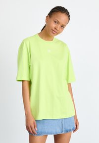 Neon zelené oversized tričko s krátkymi rukávmi a okrúhlym výstrihom, na ktorom je malé biele logo na prsiach, v kombinácii s džínsovou sukňou.