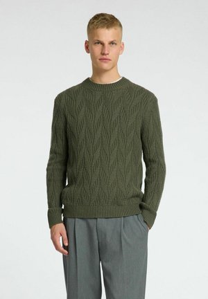 Olive green knit sweater with a textured leaf pattern, round neckline, and ribbed cuffs and hem, paired with gray trousers.