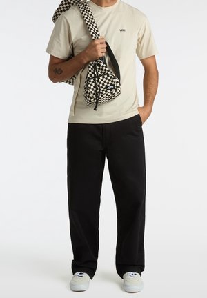 Uomo che indossa una maglietta beige Vans, pantaloni neri, sneakers bianche, mentre porta borse a scacchi bianchi e neri sulla spalla e in mano.