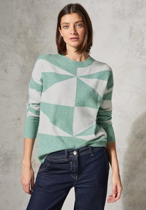 Pull en motif géométrique menthe verte et gris clair avec un col rond et des poignets côtelés, porté sur un jean bleu foncé.