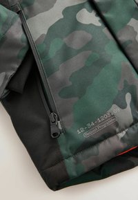 Camouflage-patterned jacket featuring a green and gray design, black accents, a zip closure, and a label with product details.