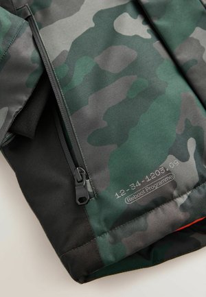 Camouflage-patterned jacket featuring a green and gray design, black accents, a zip closure, and a label with product details.