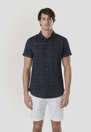 Cămașă bleumarin cu mânecă scurtă și nasturi, dintr-un material texturat, cu buzunar frontal și mâneci rulate, asortată cu pantaloni scurți albi.