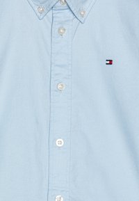 Camisa de botones azul claro, material de algodón, con un pequeño logo rojo, blanco y azul en el pecho. Presenta botones transparentes y un diseño de cuello.