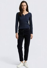 Granatowa koszulka henley z długim rękawem z ozdobnymi guzikami, zestawiona z czarnymi zwężanymi spodniami. Modelka ma na sobie białe trampki, na neutralnym tle.