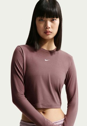 Kobieta o prostych czarnych włosach nosi długi, krótkim top o kolorze mauve z małym białym logo Nike na piersi na jednolitym tle.