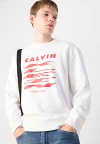Felpa bianca con grafica rossa con la scritta "CALVIN" e il logo "Calvin Klein". Presenta un colletto rotondo, polsini a costine e una vestibilità casual.