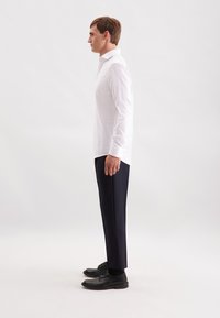 Camicia bianca a maniche lunghe con colletto a punta, abbinata a pantaloni blu scuro e scarpe eleganti nere, vista di lato.
