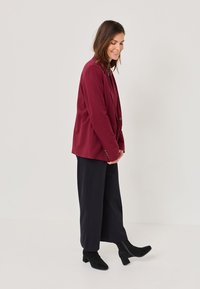 Bordo blejzer opuštenog kroja, s jednim dugmetom i suptilnom teksturom, u kombinaciji s tamnim širokim hlačama i crnim čizmama do gležnja.