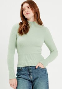 Mintgrüner Pullover mit langen Ärmeln, der über einen hohen gerippten Halsausschnitt, strukturierten Stoff und eine figurnahe Taille verfügt, kombiniert mit hellblauen Jeans.