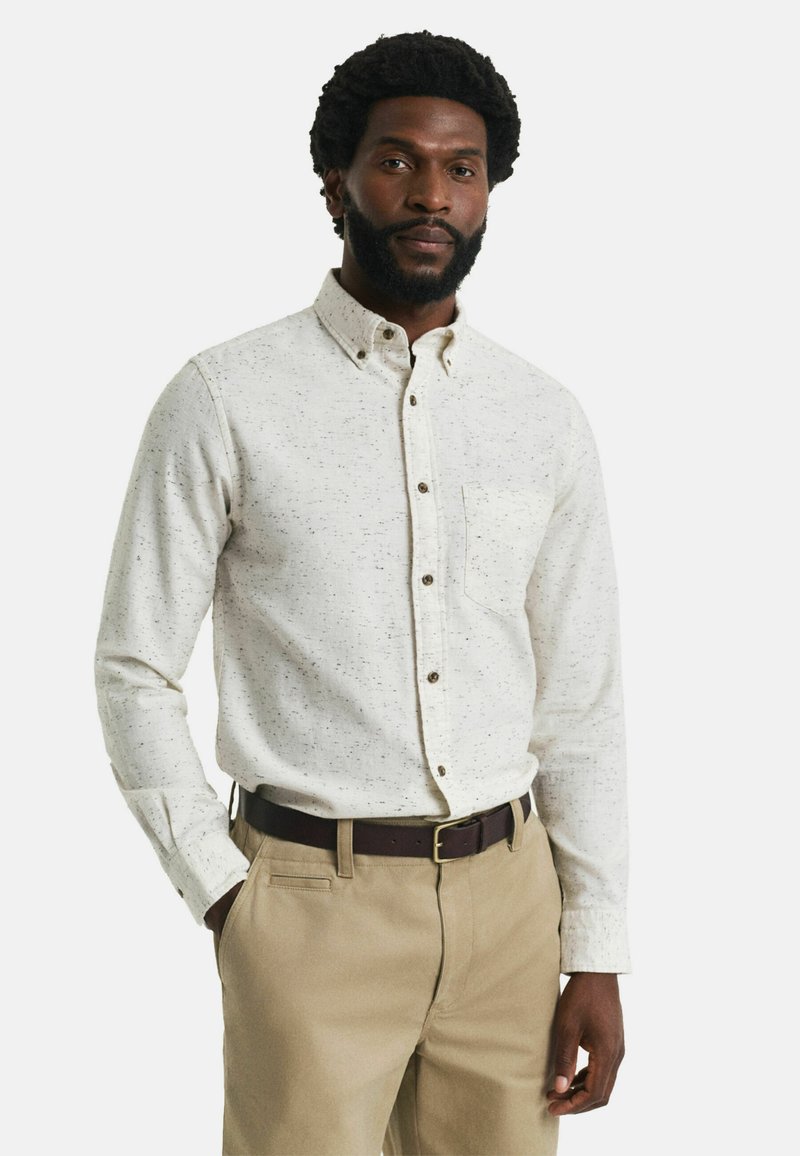 Chemise boutonnée à manches longues beige clair avec un motif légèrement moucheté, dotée d'une poche poitrine et de boutons marron, associée à un pantalon kaki.