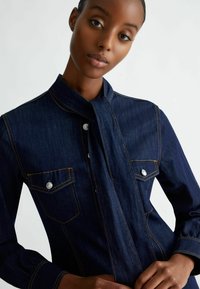 Giacca di jeans blu scuro con design senza colletto, due tasche sul petto, bottoni argentati e dettagli di cucitura gialla. Tessuto liscio e vestibilità su misura.