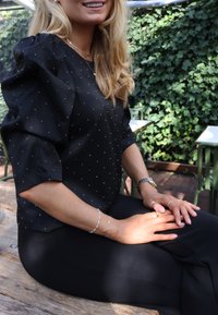 Čierna blúzka s krátkym rukávom a striebornými bodkami, v kombinácii s čiernymi nohavicami. Modelka má na sebe strieborné šperky a sedí pri drevenom stole v zelenom prostredí.