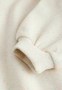 Tessuto di maglione beige chiaro con un motivo macchiettato di colori pastello. Presenta polsini a coste e una superficie morbida e testurizzata.