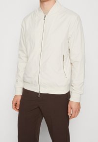 Lekka biała kurtka bomberka z przednim zamkiem błyskawicznym, ściągaczami na mankietach i u dołu. Posiada boczną kieszeń na zamek i subtelnie fakturowaną tkaninę.