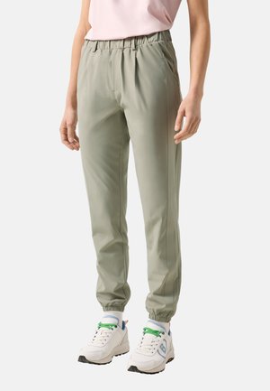 Osoba ubrana w jasnozielone spodnie dresowe z elastycznym pasem i białe trampki z zielonymi sznurówkami na jednolitym tle.