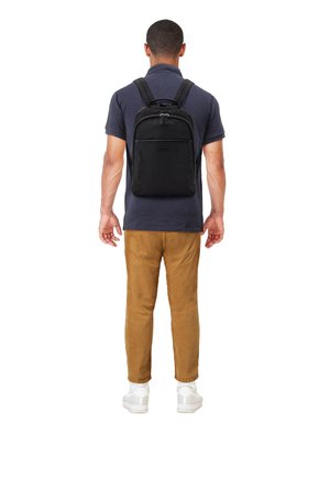Mann, der mit dem Rücken zur Kamera steht, trägt ein dunkelblaues Poloshirt, braune Hose, weiße Sneakers und einen schwarzen Rucksack mit zwei Reißverschlussfächern.