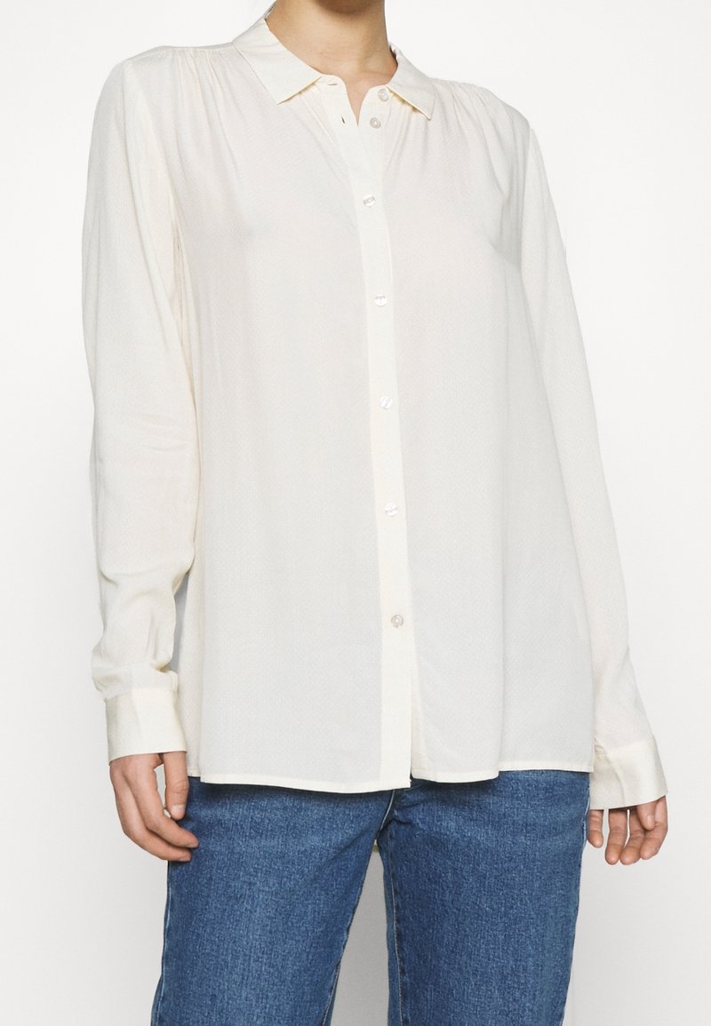 Blusa a maniche lunghe color crema con colletto, chiusura frontale a bottoni e tessuto testurizzato. Presenta polsini standard e orlo dritto.