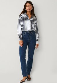 Gestreepte lange mouwen shirt in wit en marineblauw, met een knoopsluiting aan de voorkant. Hoogoplopende blauwe jeans met een rechte pasvorm. Zwarte sandaaltjes met hak.