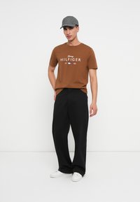 Muž v šedé čepici, hnědém tričku Tommy Hilfiger, černých širokých kalhotách a bílých teniskách, stojící na jednobarevném bílém pozadí.