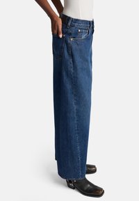 Niebieskie dżinsy o szerokich nogawkach wykonane z denimu, z wysokim stanem, tylnymi kieszeniami i widocznymi szwami, zestawione z czarnym obuwiem.